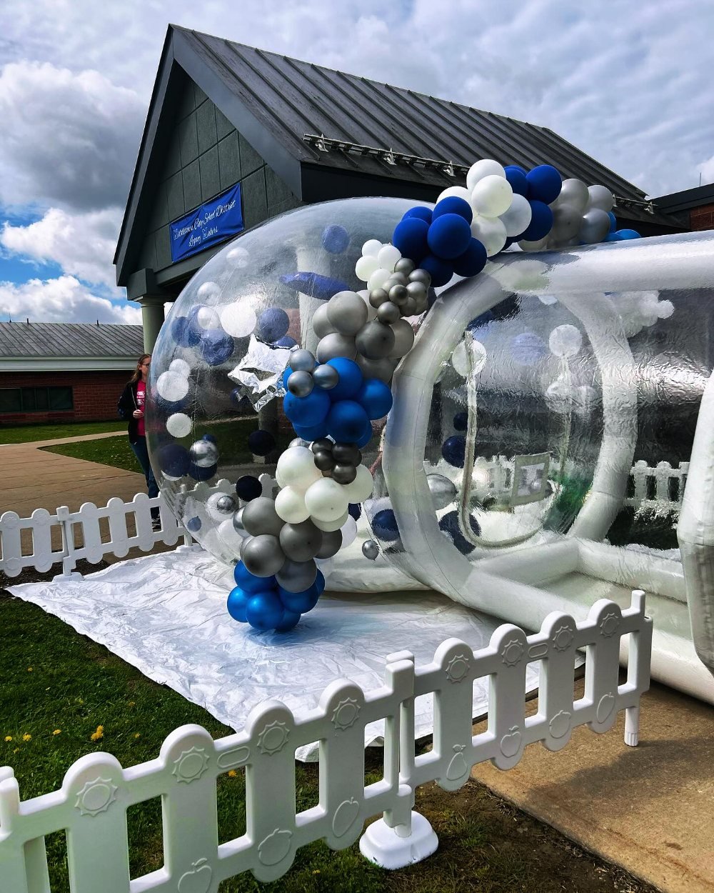 A clear bubble-dome inflatable with a navy and silver balloon arch, set up outdoors on grass behind a white picket fence.