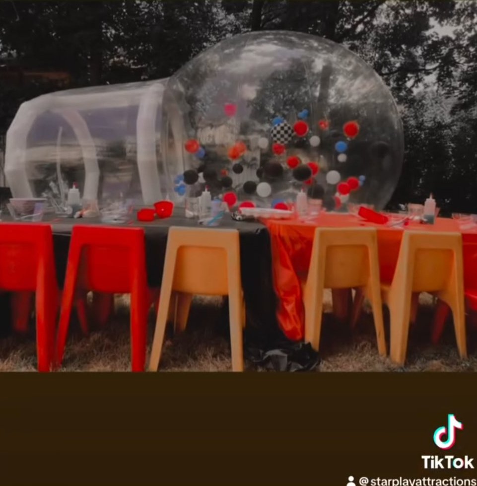 A backyard party setup with a clear bubble dome, slime tables and kids chairs arranged on the lawn.